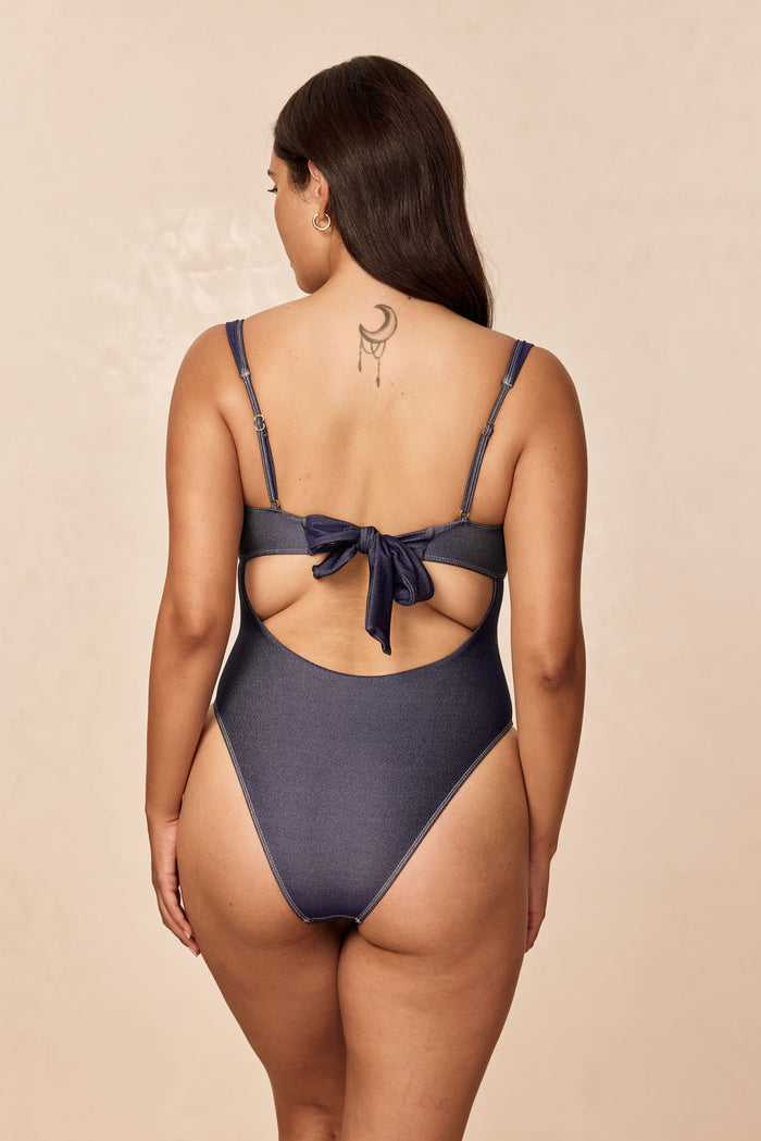 Rear view of Clovelly One Piece in Blue Denim with adjustable straps, tied back bow and high-cut leg on beige background