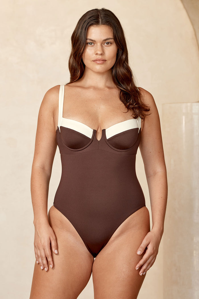 Model wearing brown one-piece swimsuit with cream underwire cups, standing front-facing against neutral beige background.