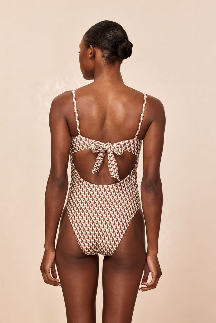 Woman seen from back wearing brown geometric-pattern one-piece swimsuit with high-cut legs and tied bow at back