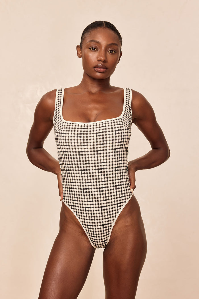 Woman wearing black-and-white gingham one-piece swimsuit with square neckline, high-cut leg, hands on hips.