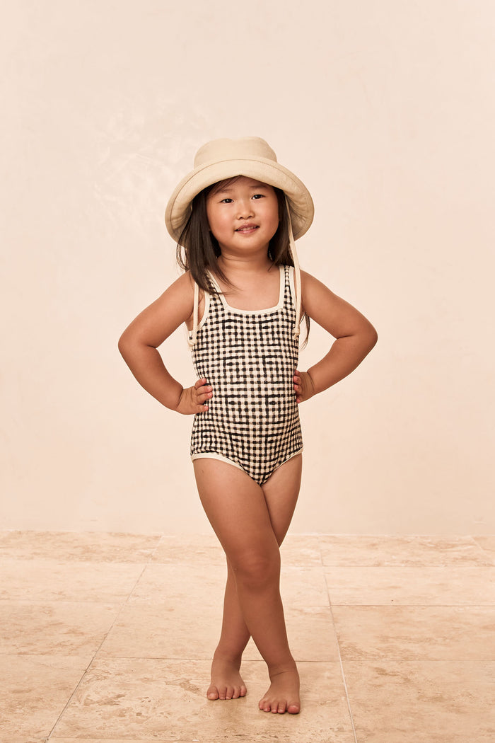 Front view of Corfu One Piece Kids in black gingham showing square neck and contrast binding on tile floor