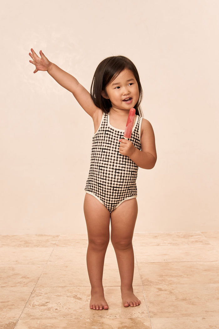Front view of Corfu One Piece Kids in black gingham showing square neckline and contrast binding on beige tile floor