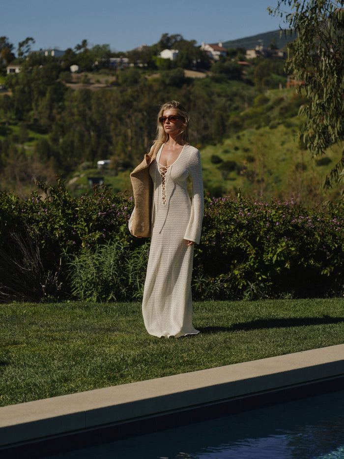 Front view of Corossol Dress in ivory lace crochet, lace-up V-neck, bell sleeves, holding straw tote by pool