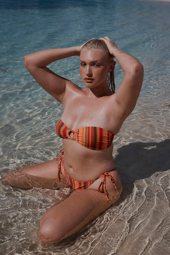 Kneeling model wearing Sardinia Stripe orange bandeau top with ring and matching tie-side bottoms in shallow water