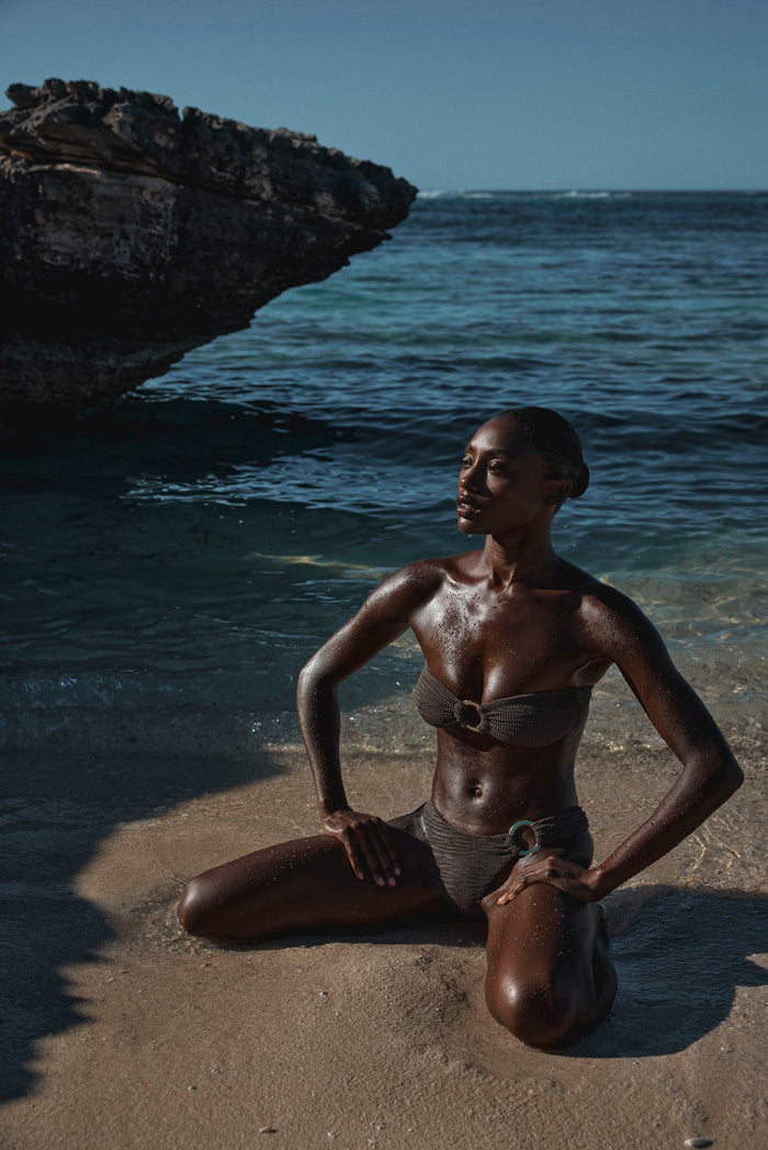 Front three-quarter view of Costa Rica top in stone crinkle showcasing bandeau shape with center ring and matching high‑waist bottoms on a rocky beach