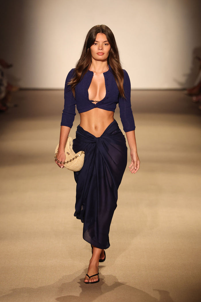 Runway model walking wearing Midnight Crinkle Crete Swim Cardi crop cardi with 3/4 sleeves, navy sarong, and straw clutch