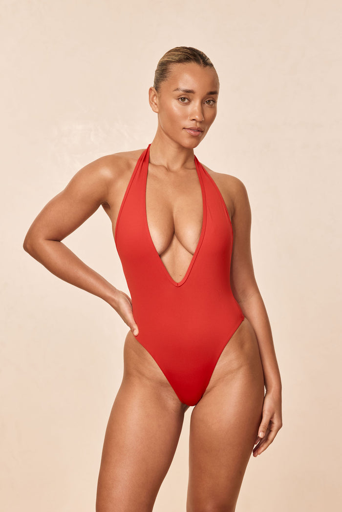 Front view of Cyprus One Piece in Chili Pepper showing deep halter plunge and high-cut leg on model against beige background