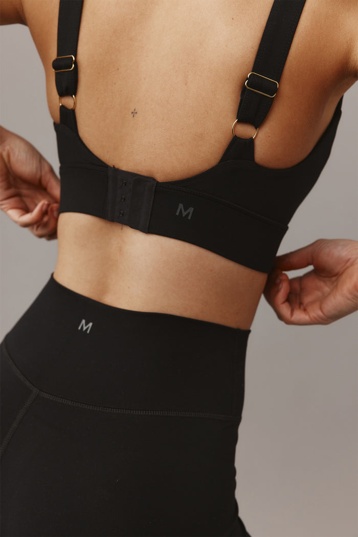 Model's back adjusting clasp on black sports bra and high-waisted leggings; small M logos on bra band and waistband.
