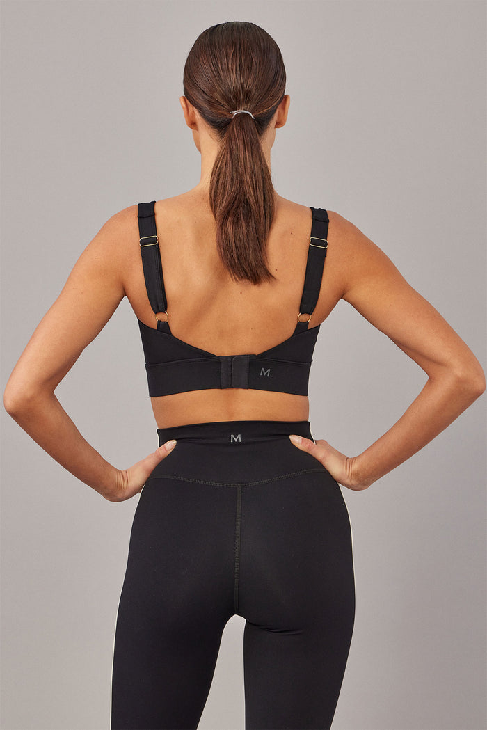 Back view of Devin Bra in black showing adjustable gold sliders, hook-and-eye closure and printed M logo on band on gray background