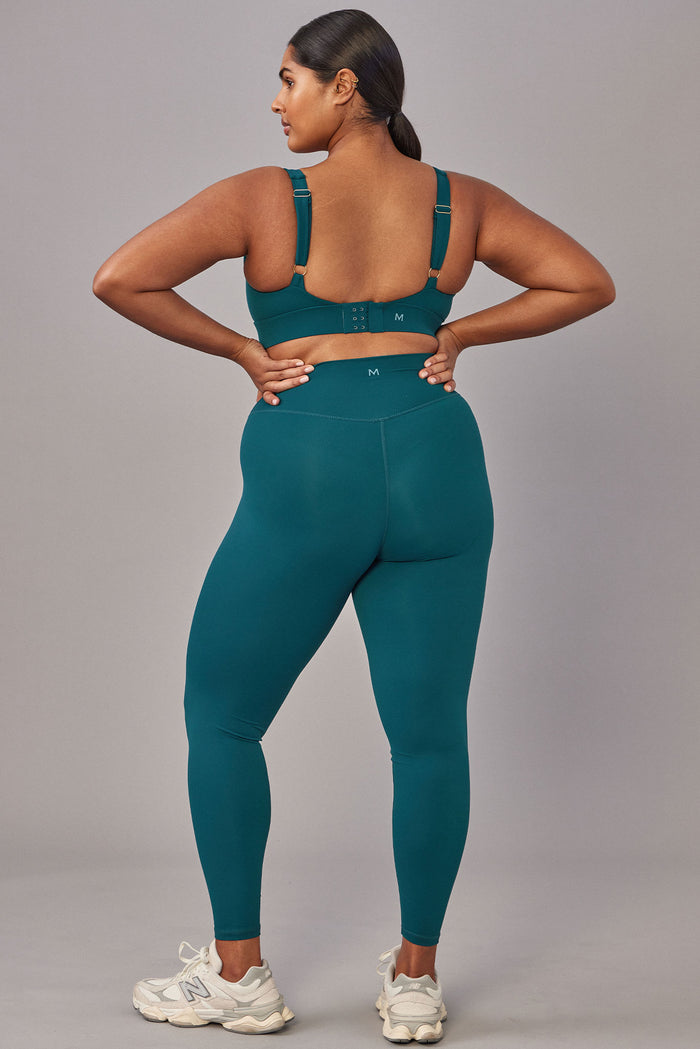 Back view of Denali The Devin Bra in evergreen on model, showing adjustable straps and three-row hook and eye back closure on gray background