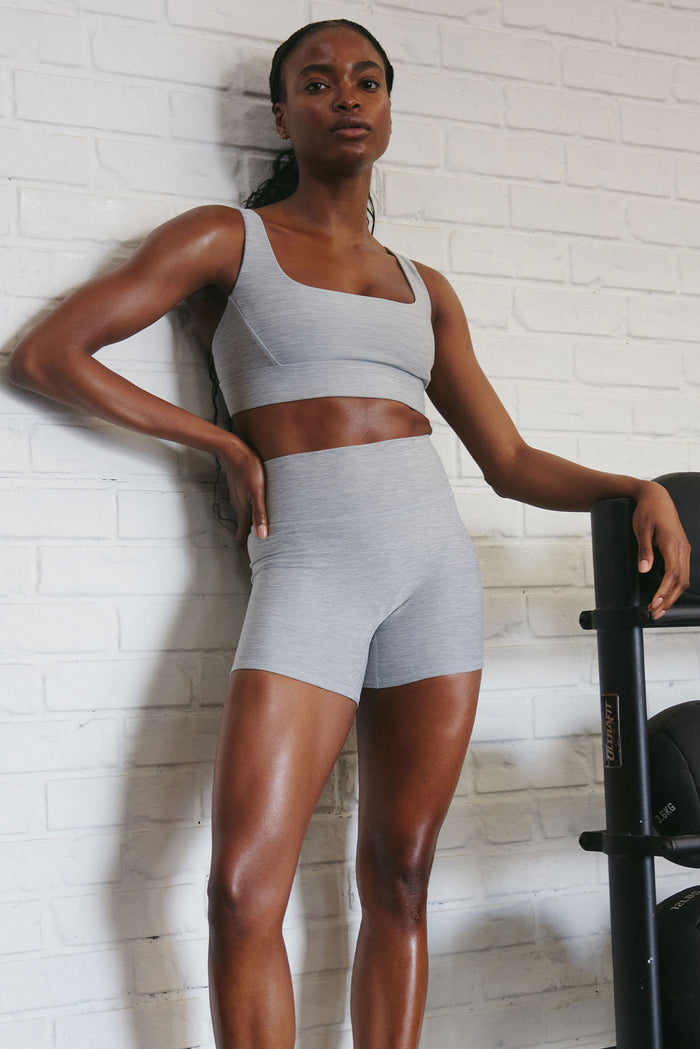 Three-quarter front view of Denali The Devin Bra in heather grey showcasing scoop neckline, thick underband and adjustable wide straps against white brick wall