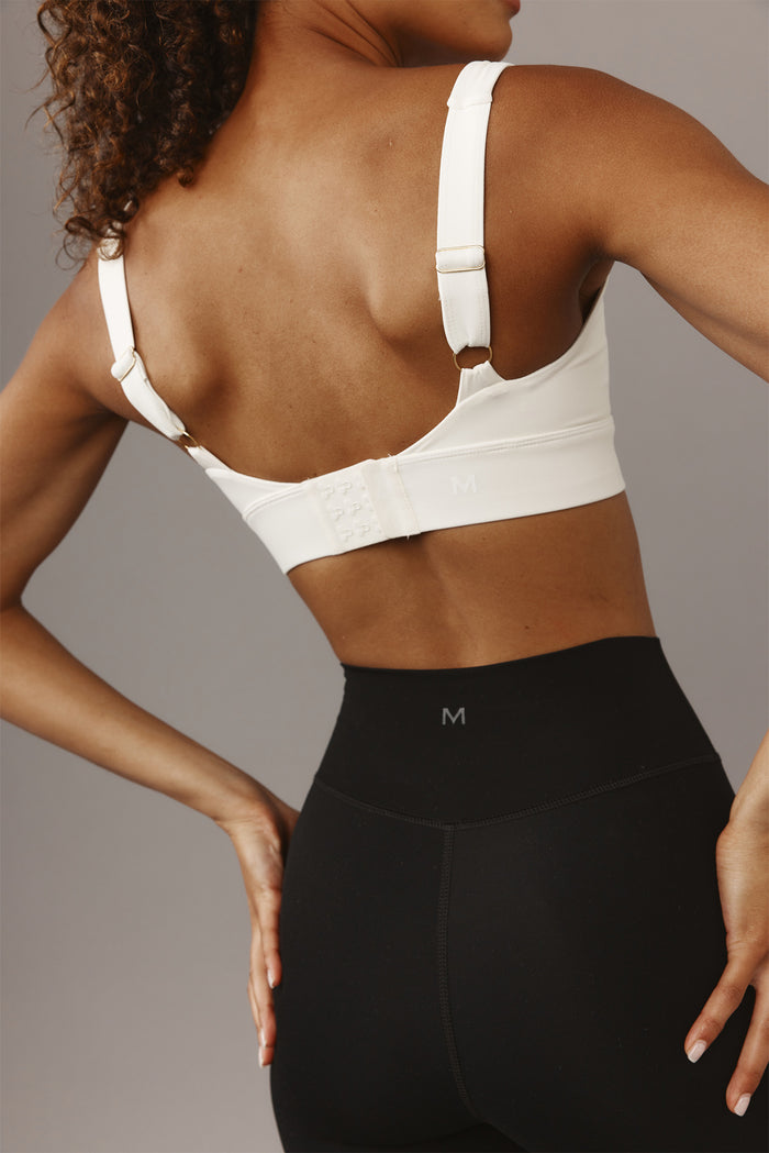 Woman's back wearing a white adjustable sports bra with hook-and-eye closure and high-waist black leggings; visible size mark 'M'.