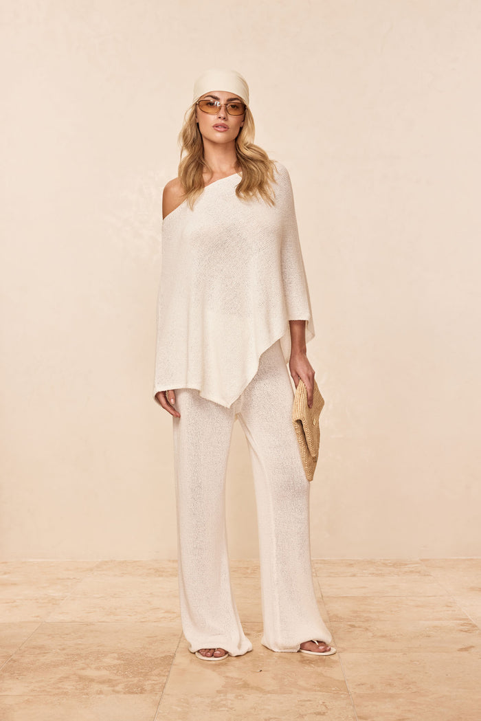 Front view of Dominica Poncho in Ivory showcasing open-knit one-shoulder drape over matching pants on beige background