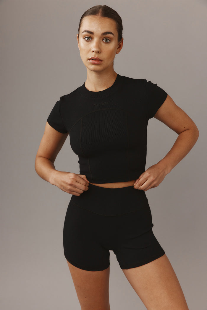 Woman wearing black ribbed crop top and high-waist shorts adjusting her waistband, faint 'MONDAY' logo on chest.