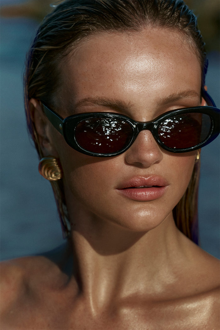 Close-up of Es Vedrá Sunglasses in black showing oval acetate frames and dark reflective lenses on a model’s face