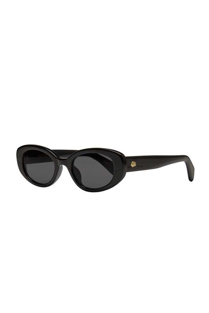 Three-quarter view of Es Vedrá sunglasses in black, cat-eye acetate frame, dark lenses, gold rivet on white