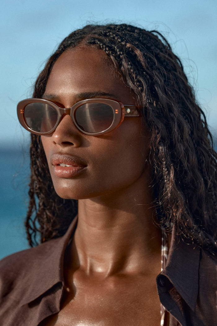 Close-up of model wearing brown oval Es Vedrá sunglasses with translucent tortoiseshell frame and sea in background