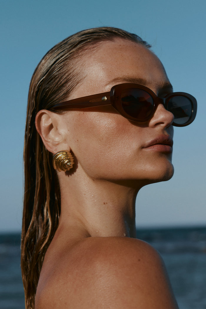 Close-up profile of Es Vedrá sunglasses on model showcasing brown translucent tortoiseshell oval frames and gold temple stud, sea background