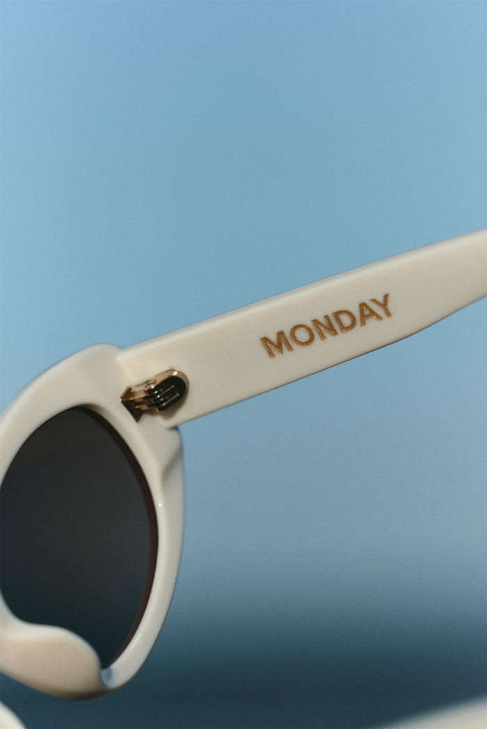Close-up of Es Vedrá sunglasses arm showing gold MONDAY lettering and metal hinge on ivory acetate frame against a blue background