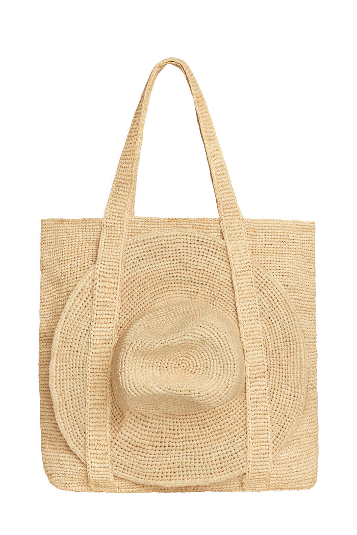 Three-quarter view of Exuma Bag in Natural showing hand-crocheted raffia texture, long shoulder straps and matching raffia hat on white background