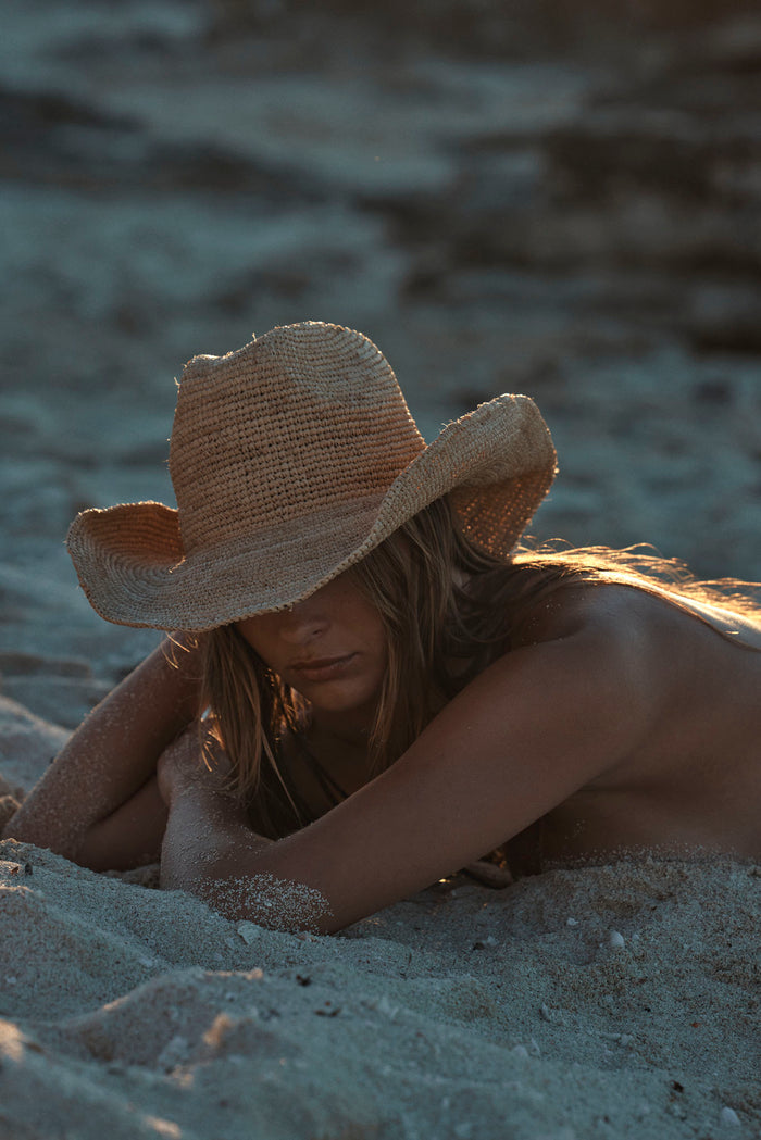 Angled view of Exuma Raffia Hat in straw on model showing shapeable brim and inner tie band on sandy beach