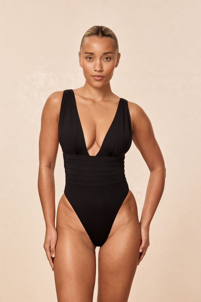 Front view of Florence one-piece swimsuit in black showcasing deep plunging V-neck, banded waist, and high-cut leg on beige background