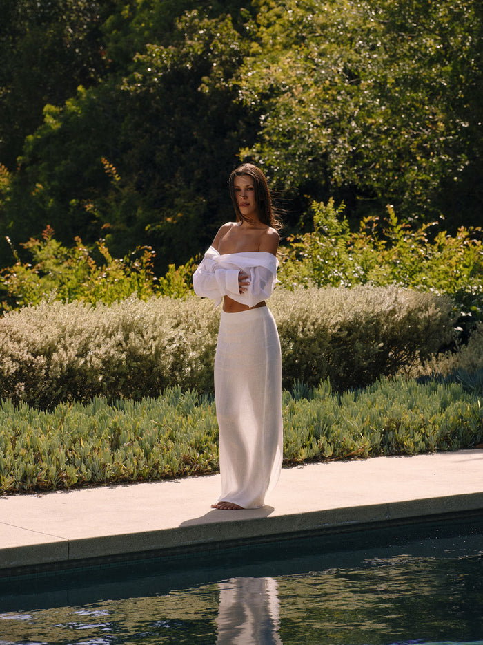 Front view of Fontelina linen maxi skirt in white showcasing high waist and slim silhouette on model by poolside with greenery