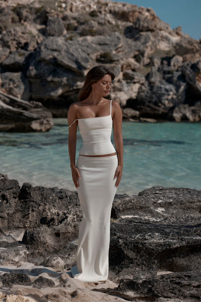 Front view of Fontelina skirt in ivory ribbed knit showcasing long fitted column silhouette with rocky beach and blue sea