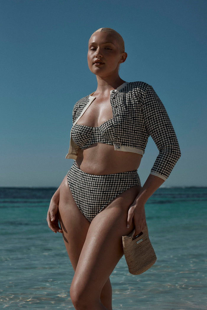 Model wearing black gingham Formentera bottom and balconette top with a cropped cardigan at the beach, holding a woven bag