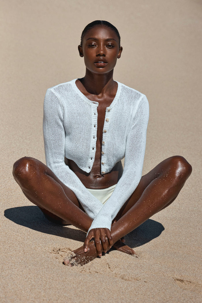 Front view of Genoa Cardigan in ivory open-knit, cropped button-front with long sleeves on model seated on sandy beach