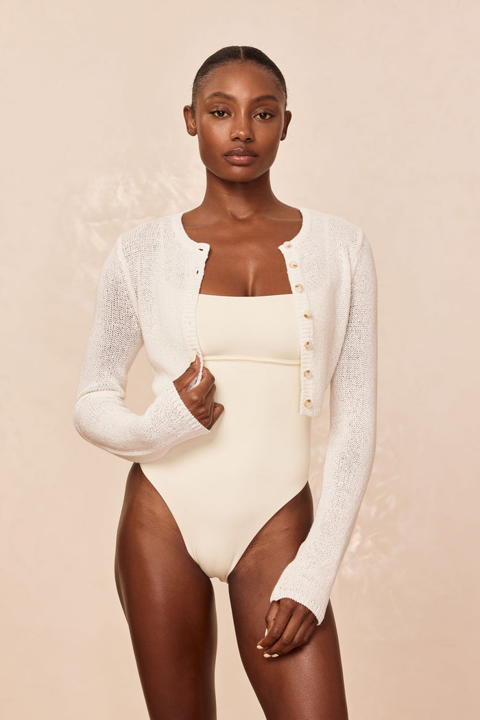 Model wearing a cream one-piece swimsuit and cropped white button-front knit cardigan against a neutral background.