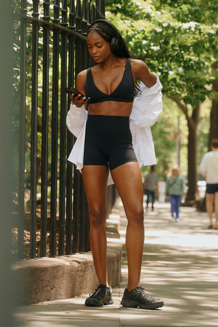 Front view of Glacier Bra in black worn by model showing V-neck and supportive band with high-waist shorts on shaded sidewalk