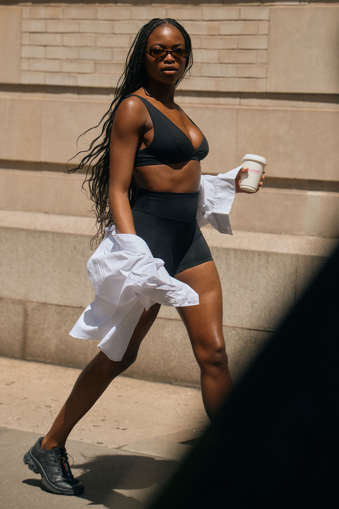 Three-quarter view of Glacier Bra in black showing V-neck and wide straps on a model walking with coffee and a white shirt