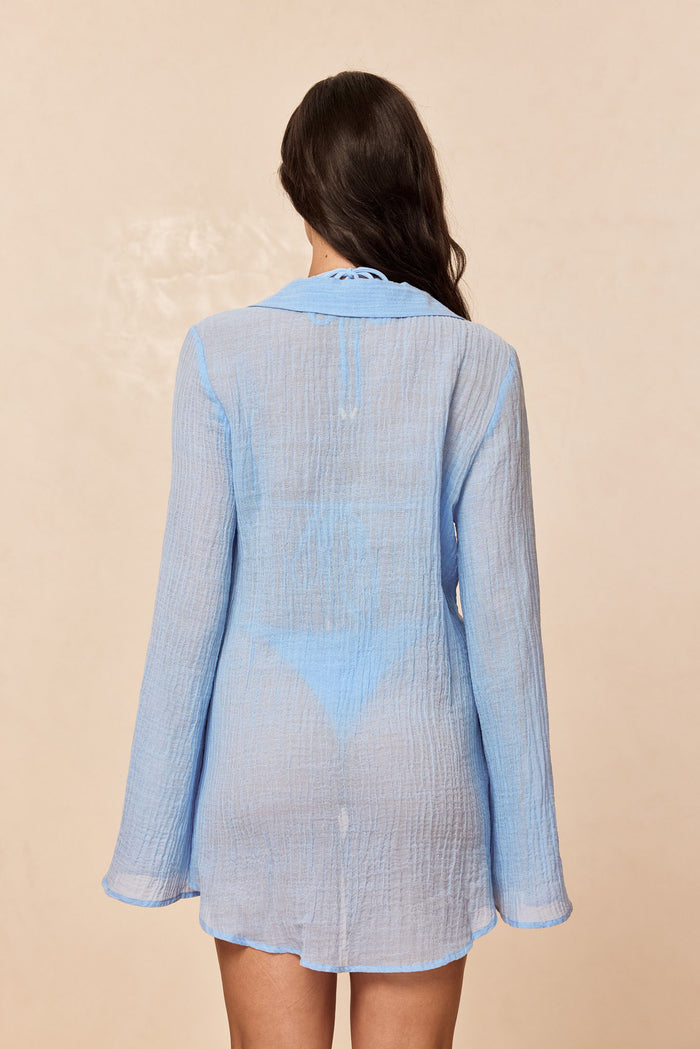 Back view of Grenada Tunic in sky crinkle linen showing sheer textured fabric, long sleeves, and relaxed fit on model against beige background