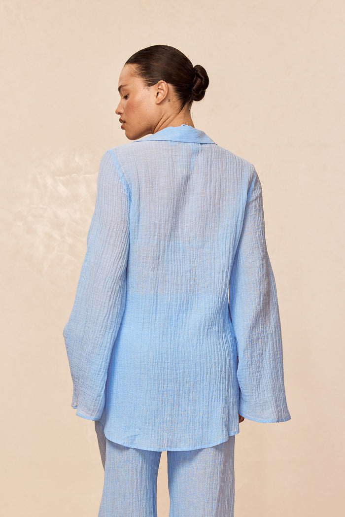 Back view of Grenada tunic in sky crinkle linen showcasing sheer crinkled texture and long bell sleeves on beige backdrop