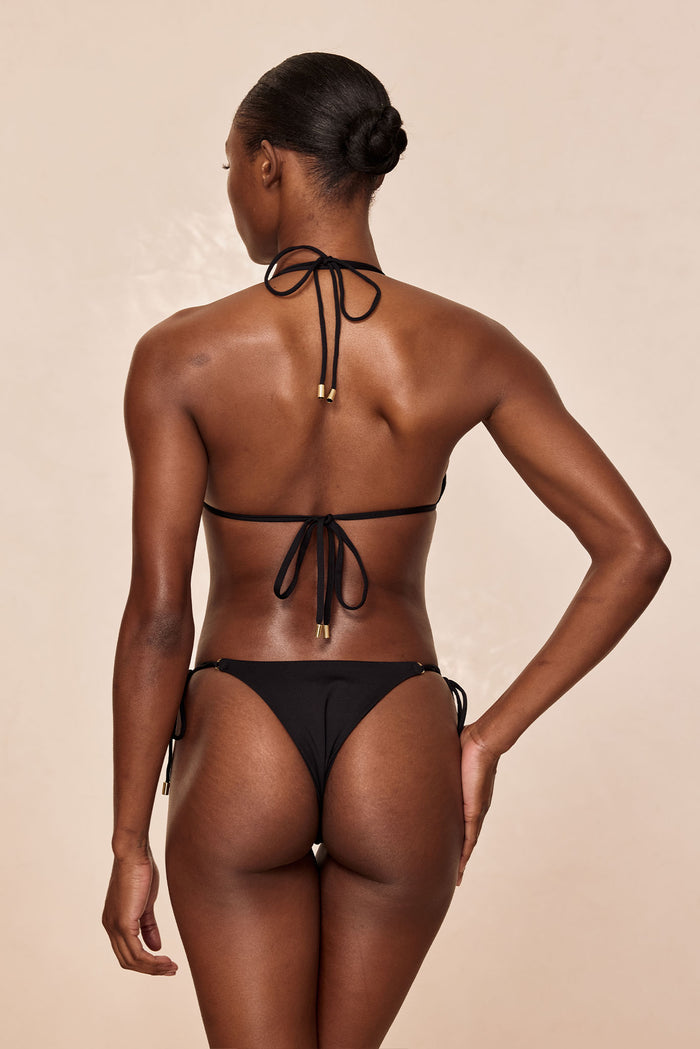 Rear view of model wearing a black tie-back halter bikini and black tie-side bottoms, hair in a low bun on beige background.