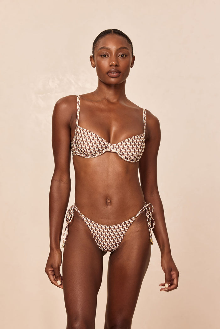 Woman wearing brown-and-cream geometric-print underwire bikini with tie-side bottoms, standing against beige backdrop