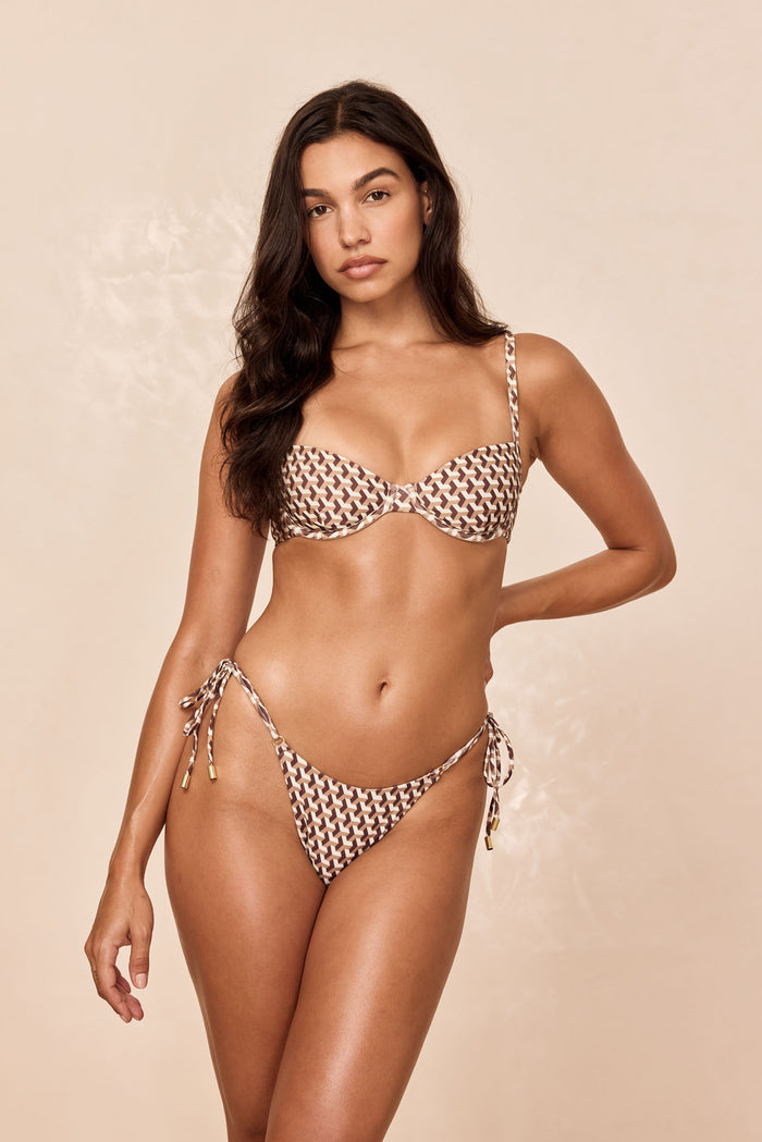 Woman modeling brown-and-cream geometric-print bikini: underwire balconette top and tie-side bottoms on beige backdrop