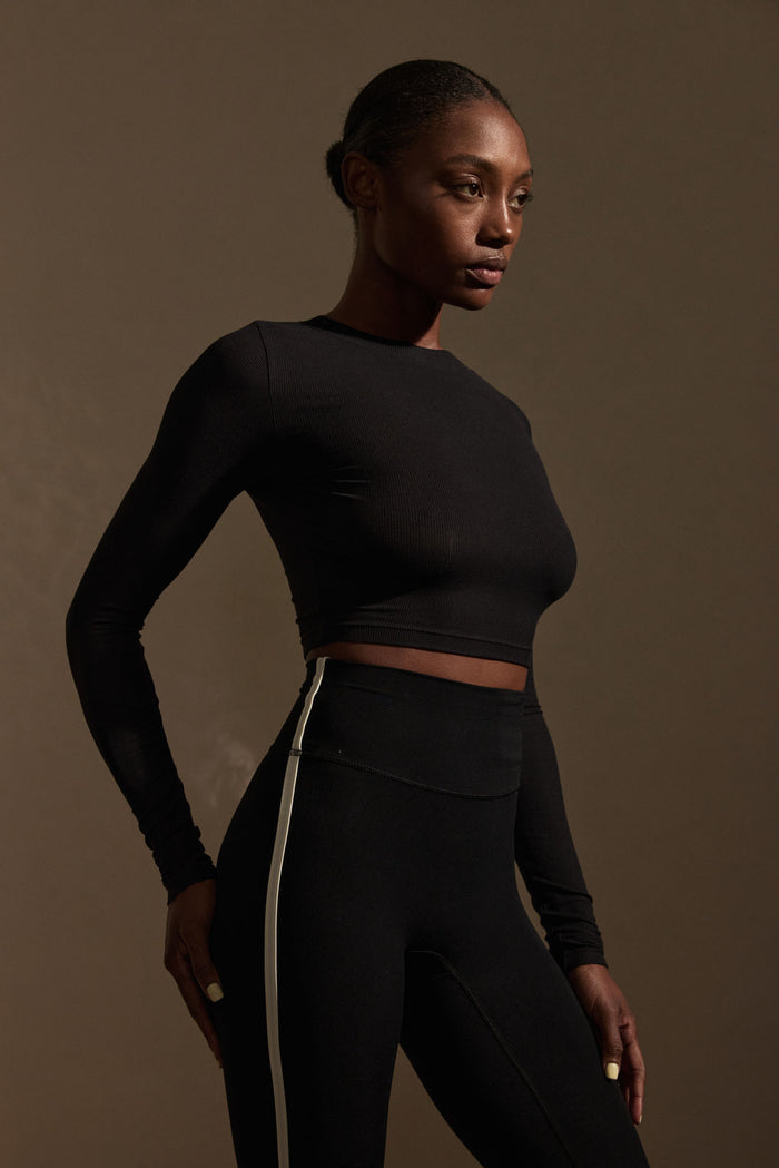 Three-quarter view of Hayworth Long Sleeve crop top in black lightweight rib showing long sleeves and cropped fit on brown studio background