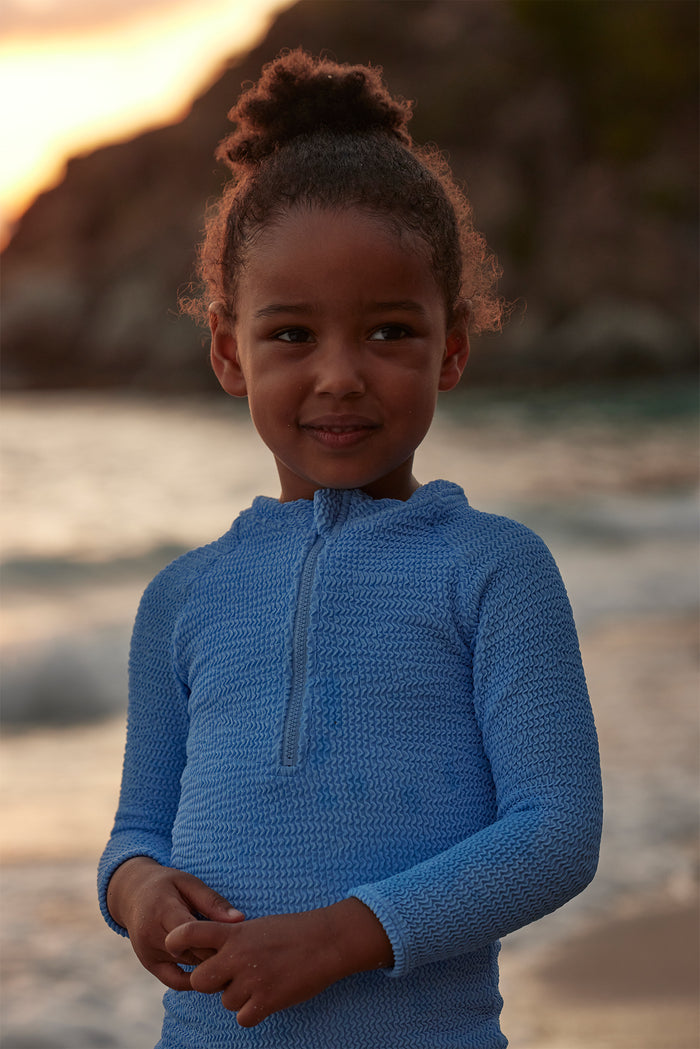 Front view of Hilo One Piece in Azure Crinkle highlighting textured crinkle fabric, center-front zip, and long sleeves on a child model standing on a beach at sunset
