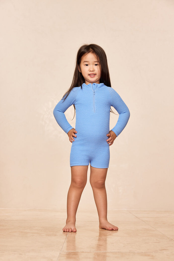 Front view of Hilo One Piece in Azure Crinkle showing textured fabric, center zipper and short-leg fit on beige background