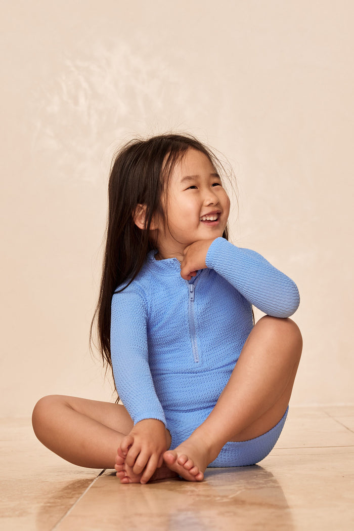 Three-quarter seated view of Hilo One Piece in Azure Crinkle showing textured fabric, long sleeves and front zip on child