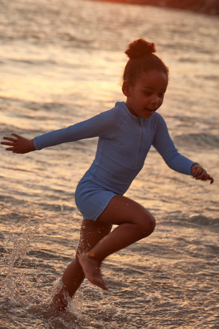 Child in blue long-sleeve swimsuit running and splashing in shallow sea water at sunset