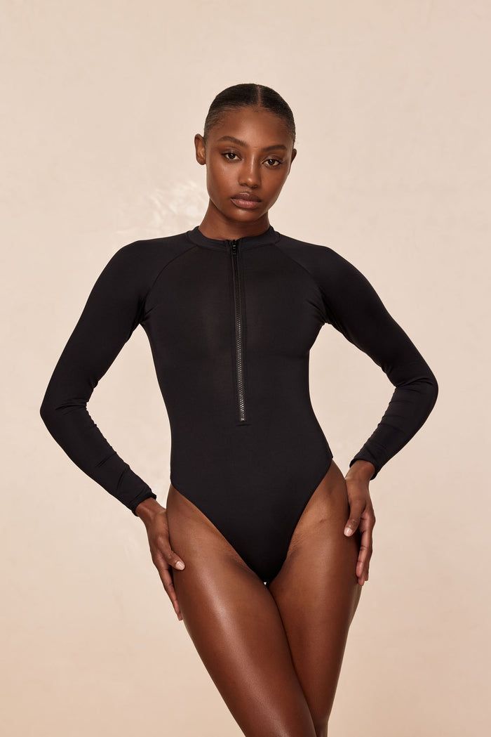 Front view of Hilo one-piece swimsuit in black on model with long sleeves, front zipper, and high-cut leg on beige background