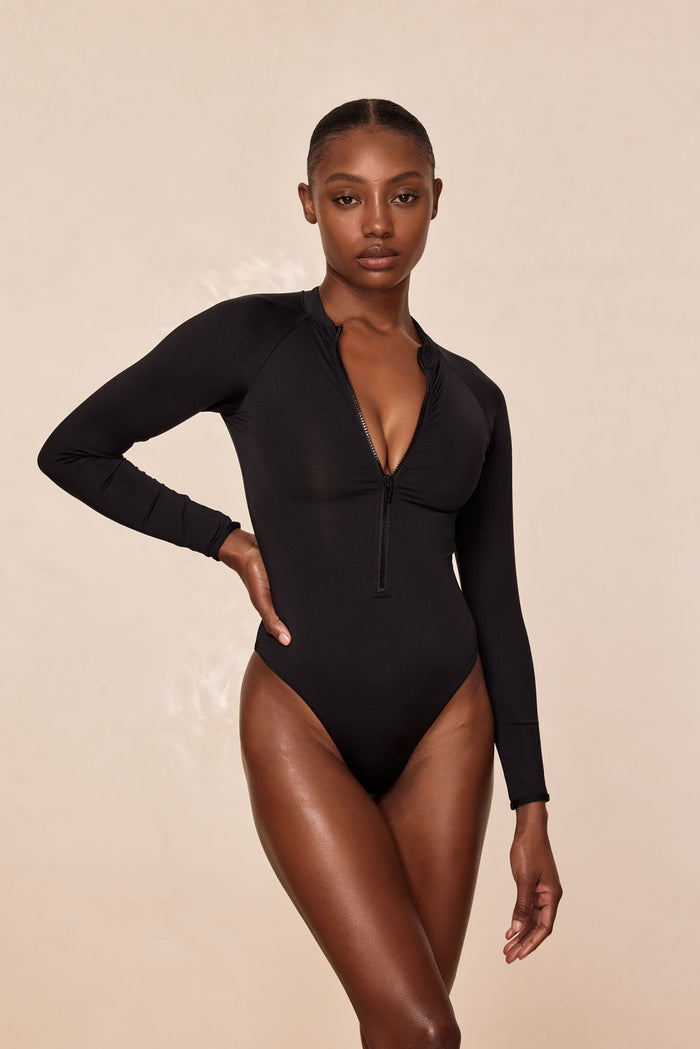Front view of Hilo One Piece swimsuit in black on model showcasing long sleeves, front zip closure and high-cut leg against a neutral beige background