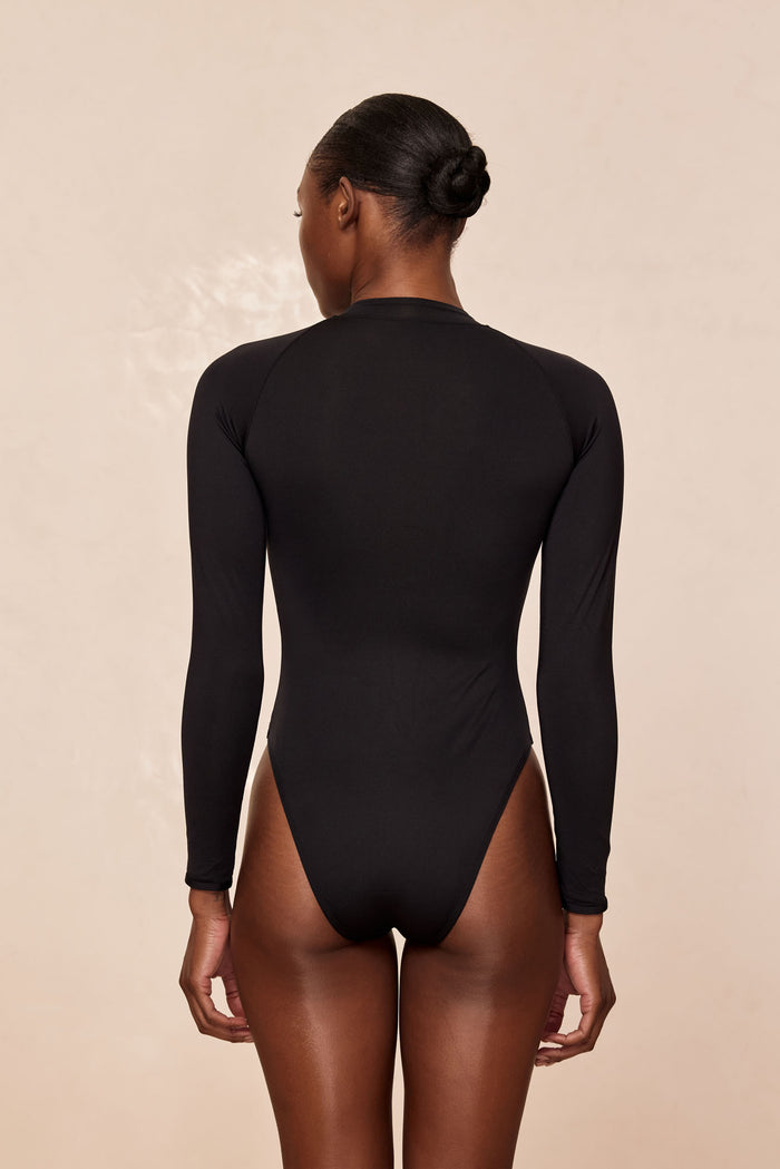 Back view of Hilo One Piece swimsuit in black showing long sleeves and high-cut leg on model against beige background