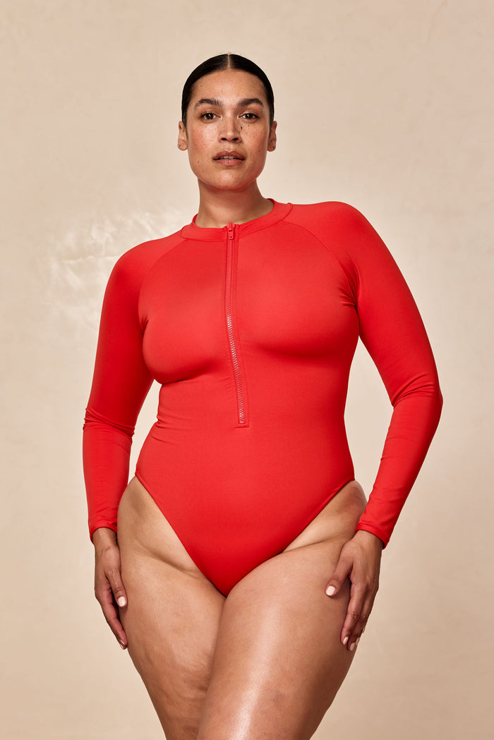 Model wearing red long-sleeve zip-front one-piece swimsuit against a beige background.