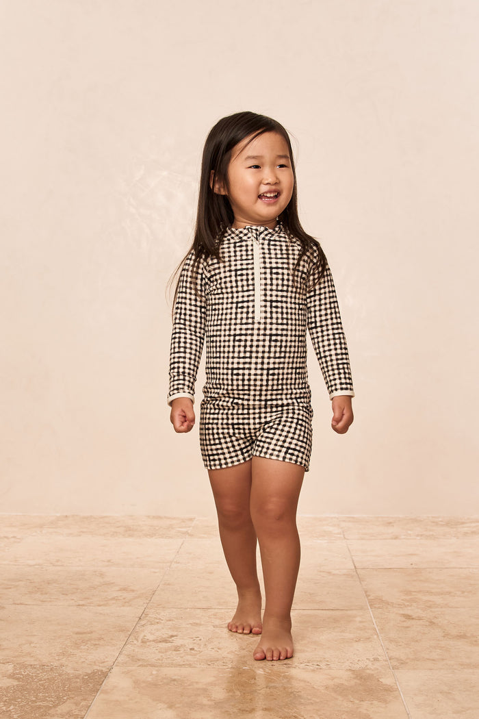 Smiling young girl walking barefoot in a black-and-white checkered long-sleeve romper with front zipper.