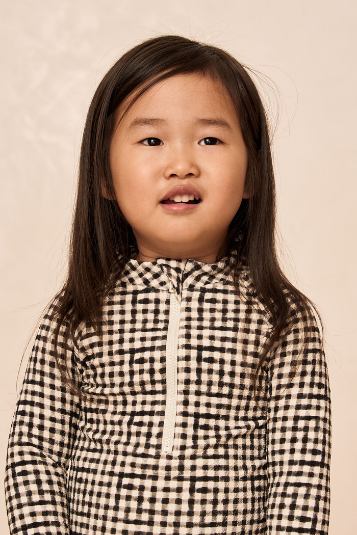 Front close-up of Hilo One Piece Kids in Black Gingham showcasing center front zipper, long sleeves, and scoop neckline on a neutral beige background