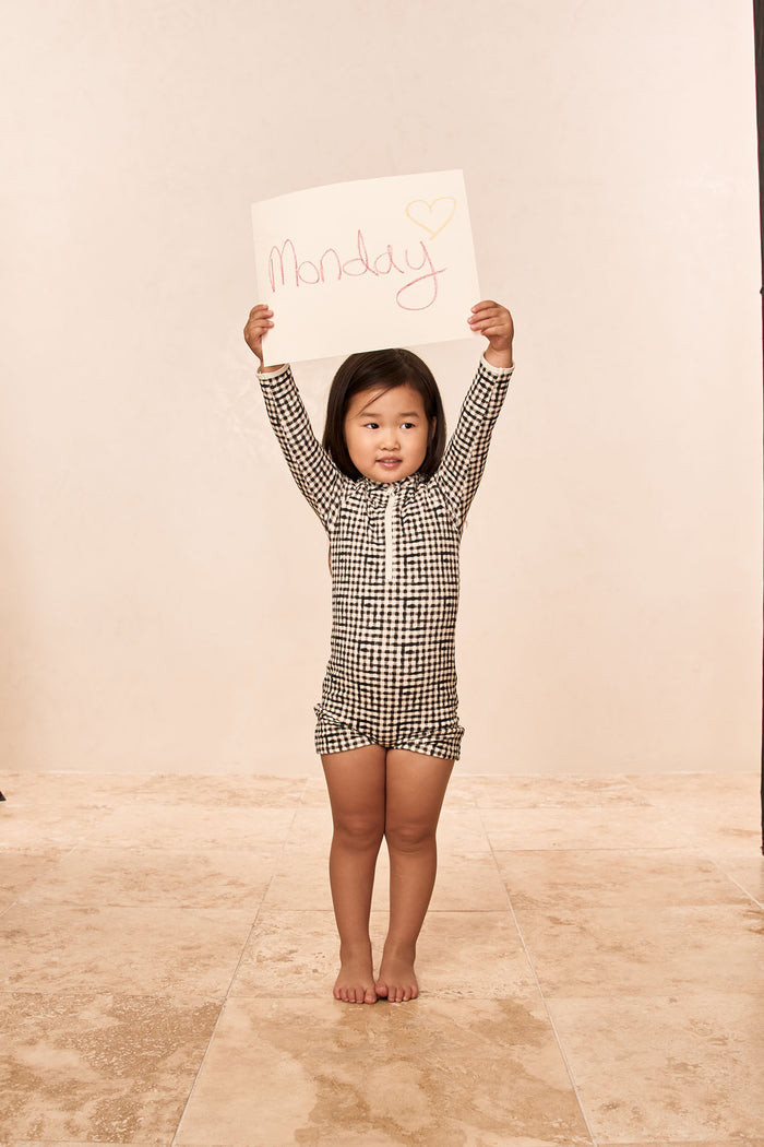 Front of Hilo One Piece Kids in Black Gingham with long sleeves and front zipper, child holding sign reading Monday